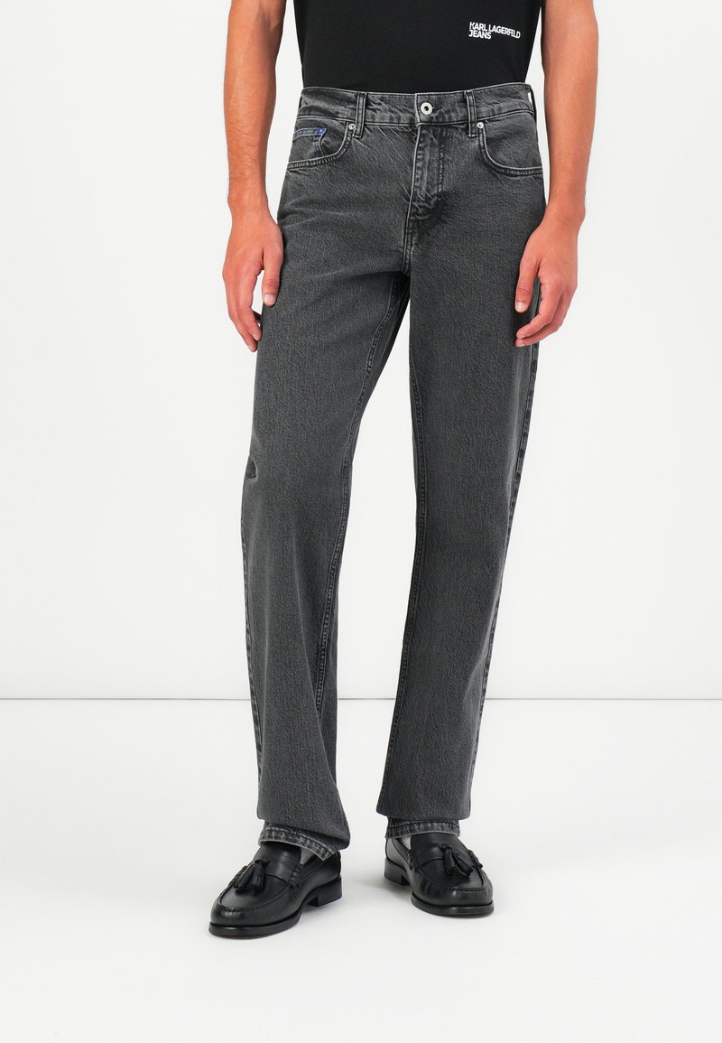 Jeans de denim negro con diseño de pierna recta, que presentan un acabado desgastado, cinco bolsillos y un cierre de botón metálico. Llevados con mocasines negros.