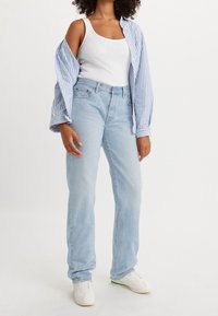 Vaqueros de tiro alto azul claro con corte recto, lavado desgastado y diseño de cinco bolsillos, combinados con una camiseta sin mangas blanca y una camisa a rayas.