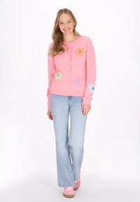 Joven sonriente, vestida con un cárdigan decorado con flores rosas, jeans de mezclilla azul claro y zapatillas rosas, de pie contra un fondo blanco.