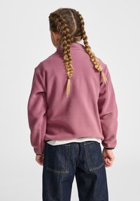 Roze fleecevest met hoge kraag en lange mouwen, gecombineerd met donkerblauwe spijkerbroek. Voorzien van gestructureerde stof en contrasterende manchetten.