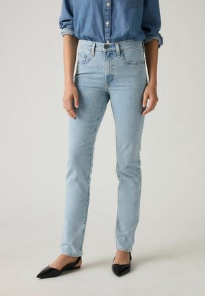 Nainen, joka pukeutunut vaaleansinisiin korkeavyötäröisiin kapeisiin farkkuihin, rullattuun farkkupaitaan ja mustiin kärjellisiin balleriinoihin, seisoo yksinkertaisen taustan edessä.