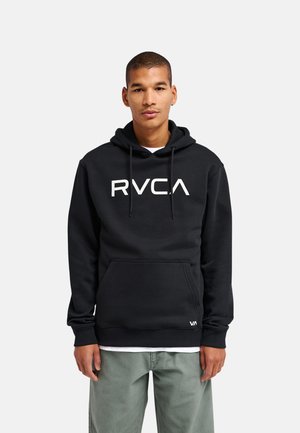 Jeune homme portant un sweat à capuche noir avec un logo RVCA blanc, pantalon vert, debout devant un fond clair uni.