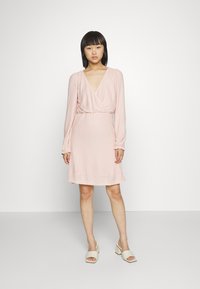 Vestido rosa claro com decote em V, mangas longas e um cós elástico. Tecido texturizado com um padrão subtil, comprimento acima do joelho.