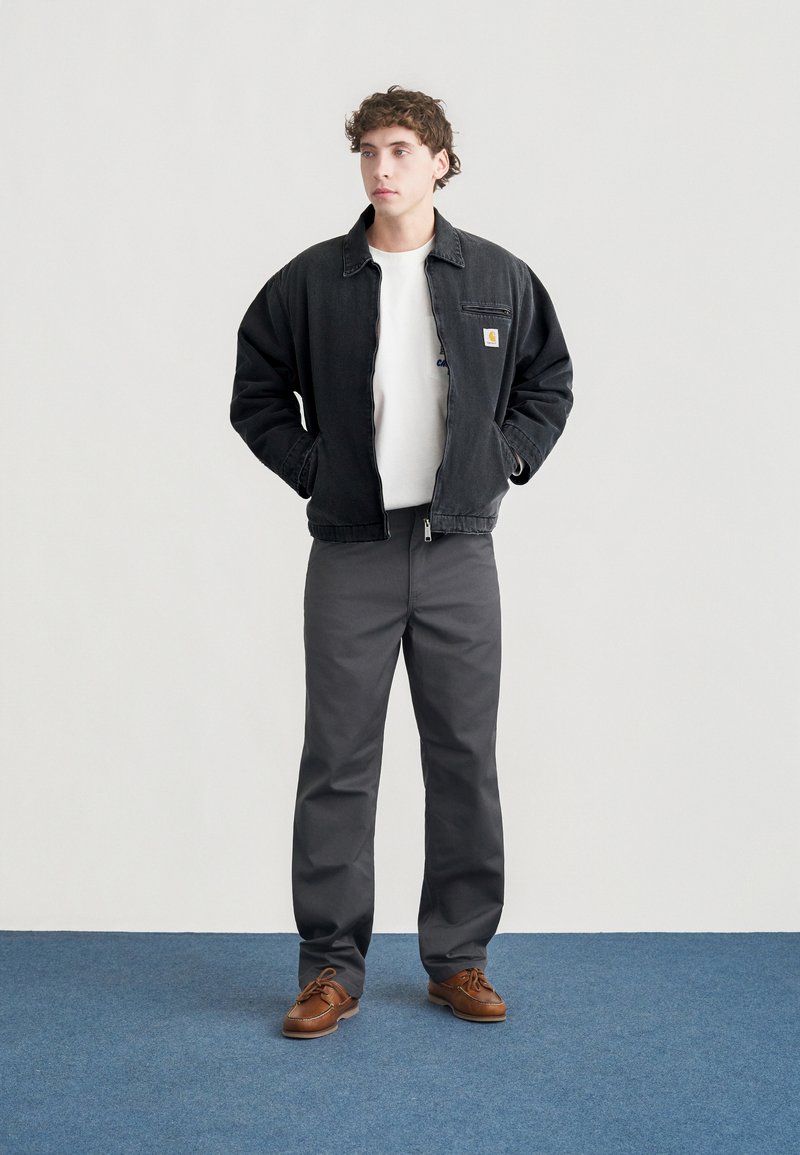 Giacca di denim nera con chiusura a zip, maglietta bianca sotto, pantaloni grigi e scarpe marroni. Il modello è in piedi davanti a uno sfondo neutro.