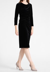 Svart sammet klänning med långa ärmar och draperad halsringning. Knälång, figurnära silhuett, slät yta, tillsammans med beige klackskor.