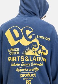 Modrá mikina s kapucňou so žltým veľkým textom: „DC SHOE CO.“ a „PARTS & LABOR.“ Grafika ruky so slnečnými lúčmi, mäkký bavlnený materiál.