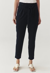 Pantaloni in morbido tessuto blu navy con pieghe, vita con laccetto, tasche laterali e orli arrotolati. Indossati con sandali bianchi minimal.