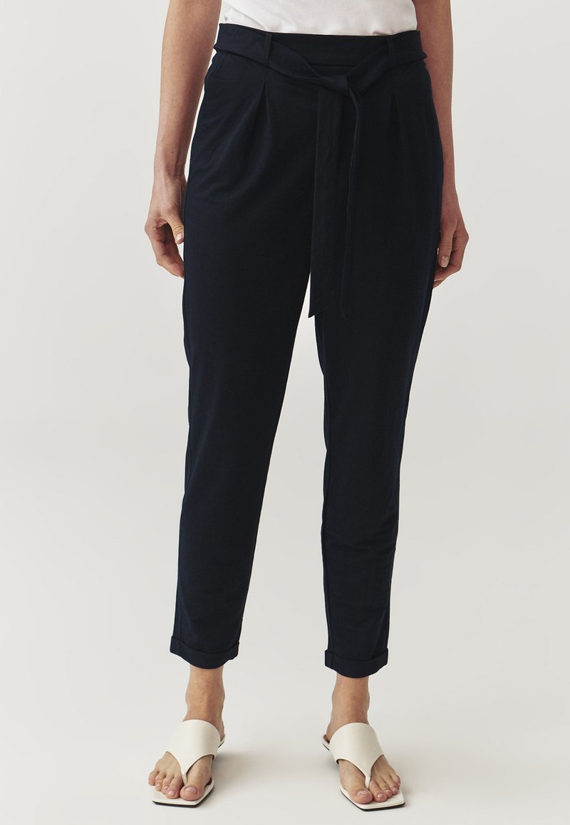 Pantaloni in morbido tessuto blu navy con pieghe, vita con laccetto, tasche laterali e orli arrotolati. Indossati con sandali bianchi minimal.