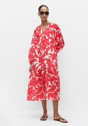 Femme portant une longue robe fluide rouge et blanche à fleurs, des lunettes de soleil foncées et des sandales marron, debout devant un fond blanc.