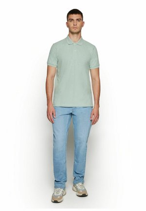 Mand stående iført en lysegrøn polo, lyseblå jeans og hvide og beige sneakers mod en ensfarvet hvid baggrund.