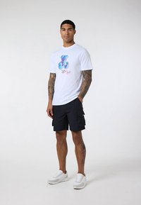 T-shirt branca de algodão com uma estampa colorida de urso, combinada com calções cargo escuros e ténis brancos, posando contra um fundo simples.