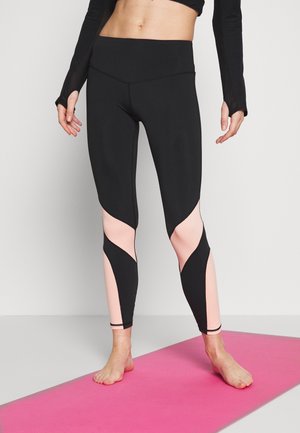 Persona che indossa leggings neri e rosa e un crop top nero a maniche lunghe, in piedi a piedi nudi su un tappetino da yoga rosa.
