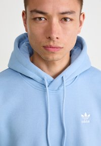 Lyseblå hettegenser laget av mykt stoff, med en stor hette, snorer og en hvit Adidas-logo på brystet.