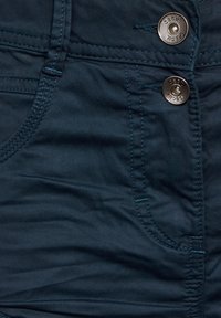 Tmavě modrozelená látka s dvěma kovovými knoflíky, kontrastním šitím a bočními kapsami. Materiál je hladký s mírným leskem.