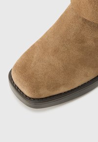 Približen posnetek rjave semiševe sprednje konice in črnega podplata škornja na svetli podlagi.