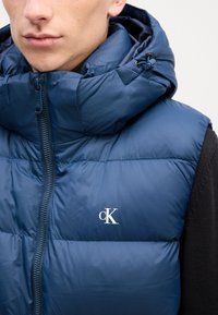 Mornarica puffer prsluk s visokim ovratnikom, patentnim zatvaračem sprijeda i detaljem s logom. Materijal izgleda sjajno, s kviltanom teksturom i podesivim rubom.