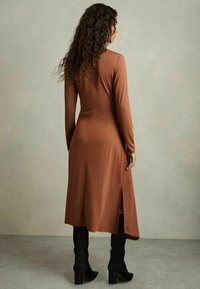 Abito lungo marrone con corsetto aderente, maniche lunghe e spacchi laterali. Indossato con stivali neri alta fino al ginocchio, tessuto strutturato e finitura liscia.
