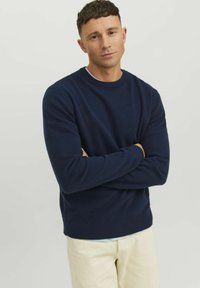 Pulover tricotat de culoare bleumarin, cu guler rotund cu tiv și mâneci lungi, purtat peste o cămașă de culoare albastră deschisă, asortat cu pantaloni de culori deschise.