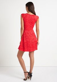 Kvinna i en ärmlös röd spetsklänning med åtsittande midja och svarta högklackade sandaler, stående vänd bort från kameran mot en vit bakgrund.