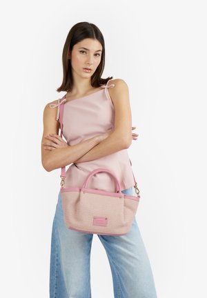 Giovane donna con capelli scuri incrocia le braccia indossando una canotta rosa e jeans azzurri, tenendo una borsa rosa con tracolla.
