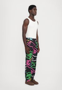 Black denim pants with colorful abstract designs in pink, green, and white. Paired with a fitted white tank top and brown shoes.