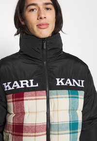 Chaqueta acolchada negra con un patrón de cuadros en rojo, beige y turquesa, con un cuello alto y un logo bordado en blanco.