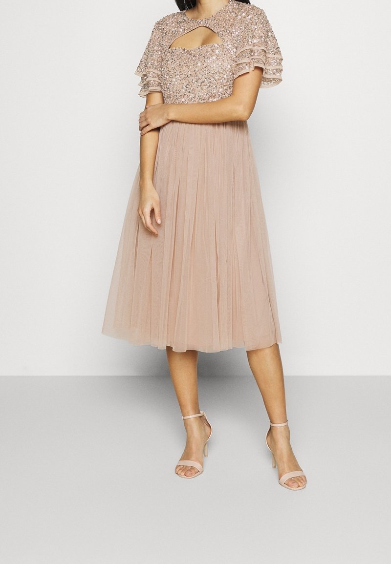 Mujer que lleva un vestido beige hasta las rodillas con un cuerpo de lentejuelas, mangas ligeras y falda de tul plisada, combinado con tacones de tira al tobillo en beige.