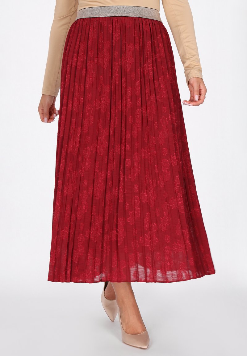 Gonna plissettata rossa con motivo floreale, vita elasticizzata con dettaglio metallico e tessuto fluido. Indossata con scarpe con tacco alto di colore chiaro.