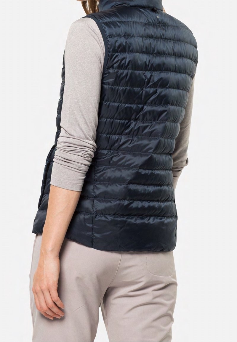 Chaleco de plumas color azul marino con un cuello alto y diseño acolchado horizontal. Cuenta con bolsillos laterales y un acabado suave y brillante. Se lleva encima de una blusa de color gris claro.