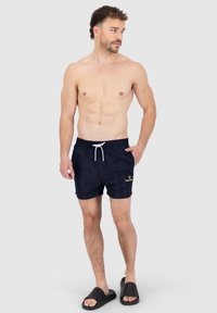 Pantaloni da bagno blu navy con vita elasticizzata e cordoncino bianco. Presentano un logo giallo nella parte anteriore. Indossati con sandali neri.