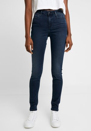 Personne portant un jean skinny bleu foncé, un top court blanc et des chaussures blanches à lacets sur un fond clair uni.