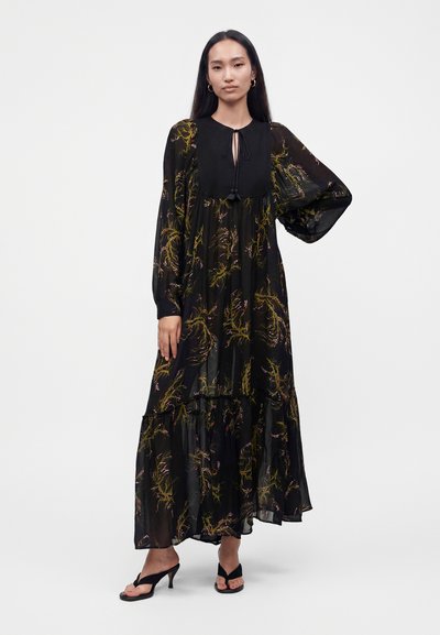 Femme portant une longue robe noire fleurie avec des manches bouffantes transparentes et des sandales noires à talons, debout sur un fond blanc.