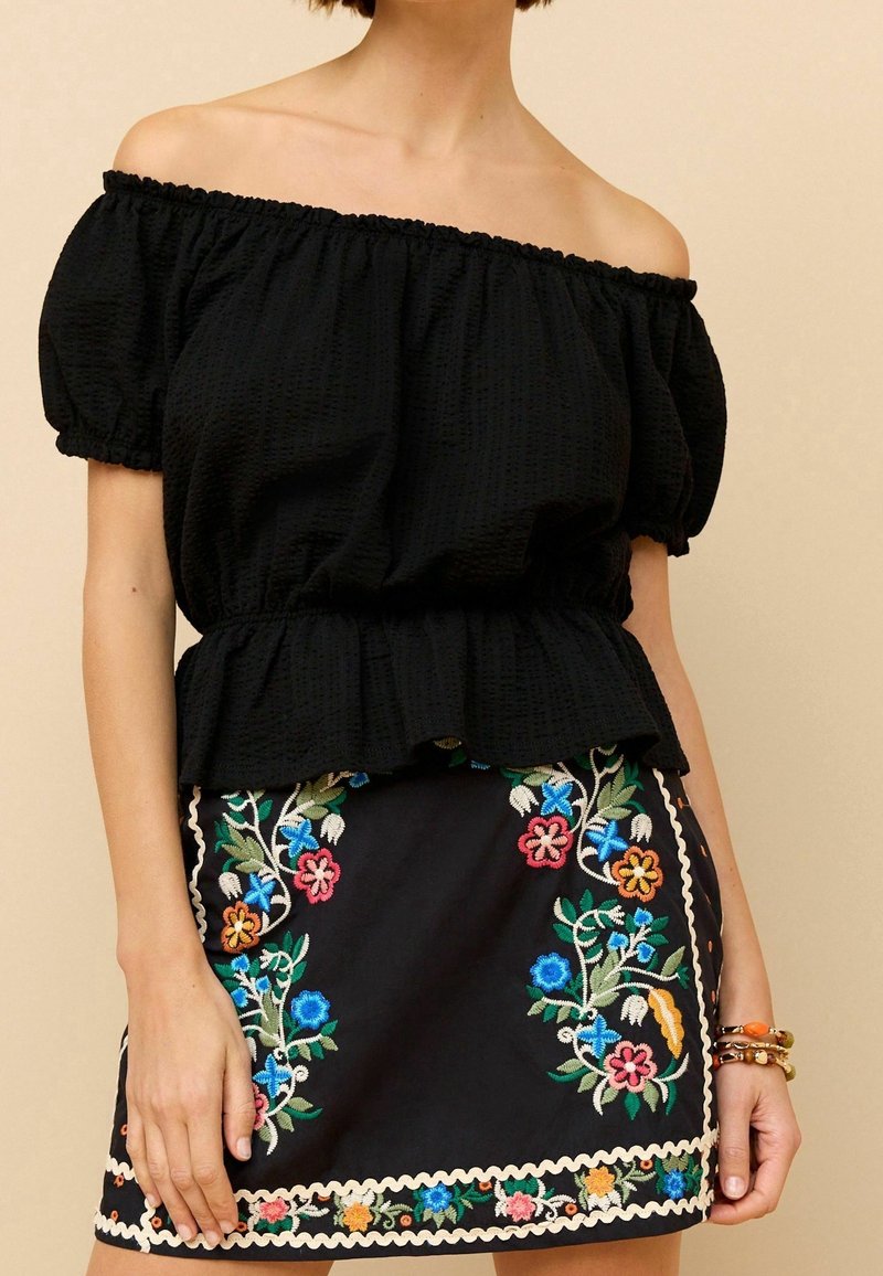 Femme portant un haut noir épaules dénudées et une jupe noire avec une broderie florale colorée à l'ourlet et sur les côtés.