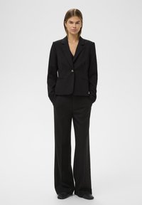 Fekete, szabott blézer egyetlen elülső gombbal, összeillő, bő szárú nadrág felett viselve. Jellemzője a zsebes rever és a klasszikus egyenes fazon.