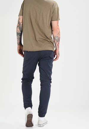 Mężczyzna stojący tyłem, ubrany w khaki t-shirt, granatowe spodnie dresowe, białe skarpetki, białe buty sportowe, z tatuażami na obu ramionach.