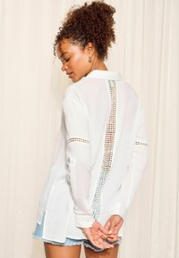 Camicia bianca a maniche lunghe con colletto, caratterizzata da un motivo a taglio lungo la schiena e le maniche. Abbinata a pantaloni corti di jeans azzurri.