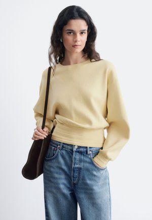 Jeune femme aux cheveux bouclés foncés portant un pull jaune pâle et un jean bleu, tenant un sac à épaule marron foncé devant un fond uni.