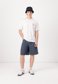 Camiseta de algodón blanca con un pequeño parche gráfico, combinada con pantalones cortos de mezclilla azul oscuro. Un sombrero tipo cubo negro y zapatillas blancas completan el conjunto.