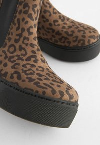 Botki platformy w leoparda z brązowej zamszowej cholewką, z czarną teksturowaną gumową podeszwą i gładkim, zaokrąglonym czubkiem.