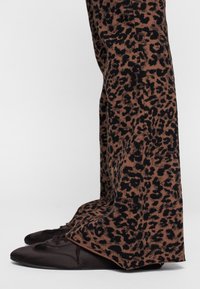 Pantalones marrones con estampado de leopardo hechos de un tejido suave, con un corte relajado y perneras acampanadas. Combinados con zapatos de cordones marrones oscuros.