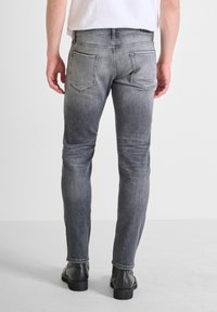 Jeans en denim gris avec une coupe slim, deux poches arrière, des coutures visibles et un léger dégradé. Portés avec des chaussures noires et une chemise blanche.