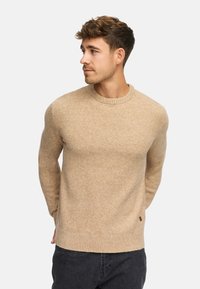 Beige wollen trui met een ronde halslijn, geribde mouwen en zoom. Heeft een subtiele textuur en een klein merkteken aan de zijkant.