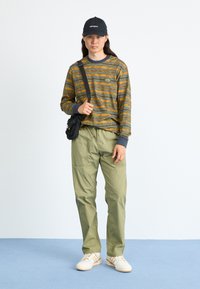 Long-sleeve striped shirt in olive green and yellow, loose olive pants, black cap, and white sneakers with light brown stripes. Black crossbody bag.