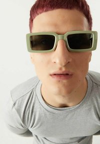 Lunettes de soleil rectangulaires vertes avec des verres teintés sombres, portées par une personne en chemise gris clair, sur fond blanc uni.