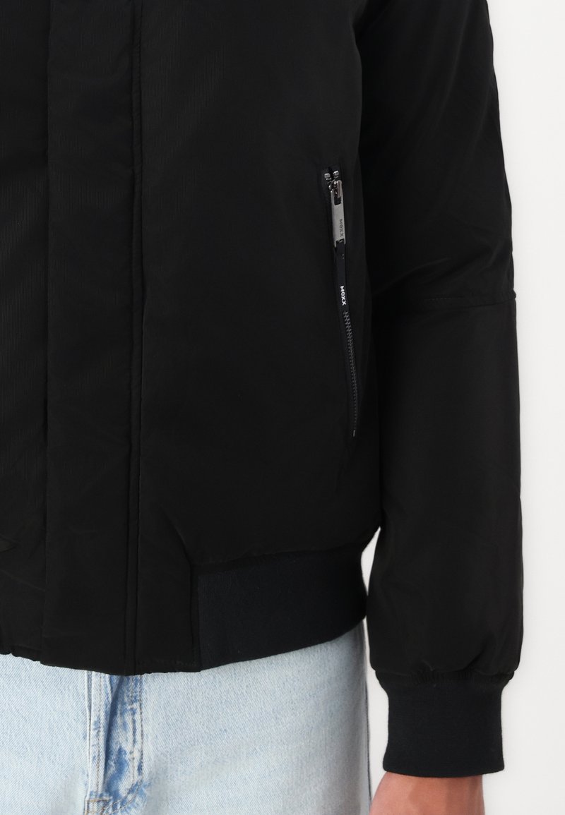 Chaqueta bomber negra con un bolsillo con cremallera, puños y dobladillo acanalados. Fabricada con un material ligero y suave que tiene un acabado ligeramente brillante.