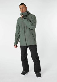 Giacca impermeabile verde con colletto alto, zip frontale, molteplici tasche con zip e pantaloni neri. Il modello indossa scarpe nere e bianche.