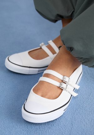 White Mary Jane-style sneakers with double buckle straps worn with green pants on a blue carpeted floor.