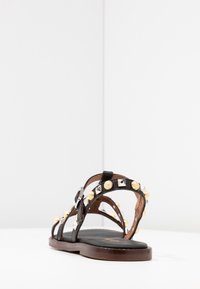 Sandal de cuero negro con acentos de tachuelas doradas y plateadas, correa ajustable, suela de madera plana y diseño de punta abierta.