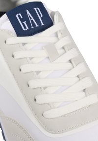 Weiße Sneaker mit grauen Wildlederelementen, strukturiertem Stoff, flachen weißen Schnürsenkeln und einem marineblauen Tab mit dem "GAP"-Logo.