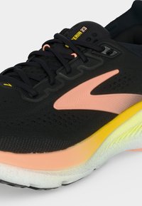 Chaussure de running noire avec logo pêche, lacets noirs, accent jaune sur la semelle intermédiaire et étiquette portant la mention "BERIN 23" sur la languette.